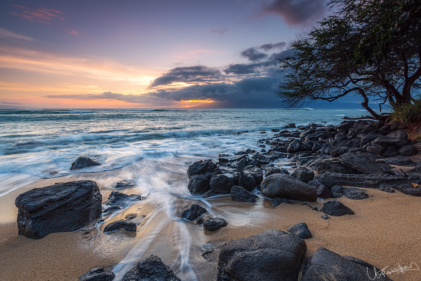 Beach scene with rocks, ocean waves, and a tree at sunset.