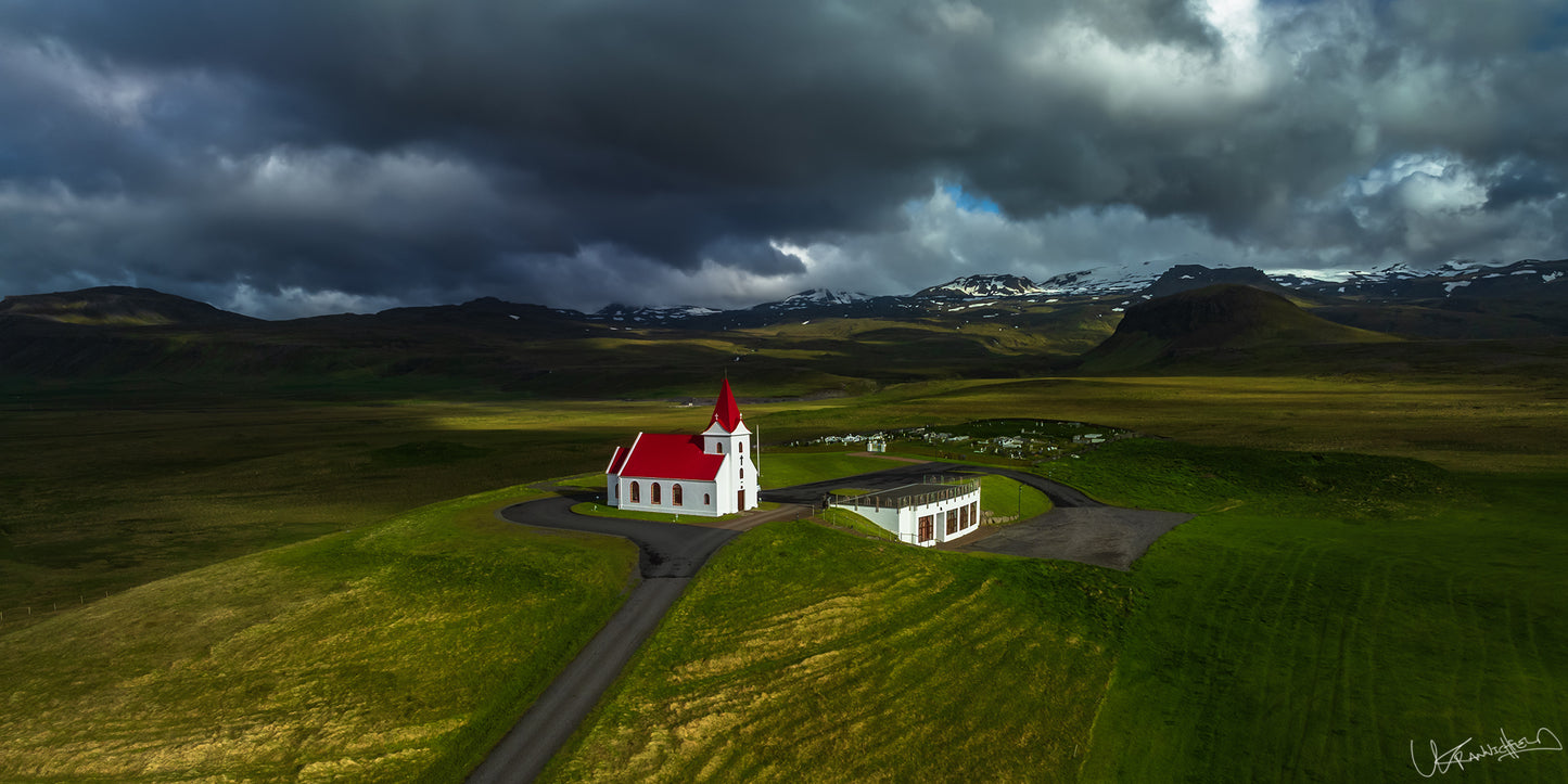 White church with a red roof in a green landscape under dark, stormy skies.