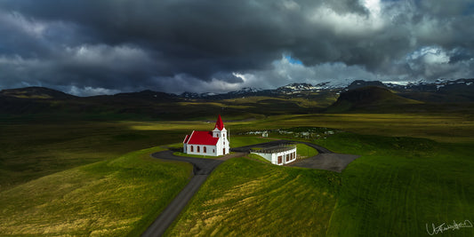 White church with a red roof in a green landscape under dark, stormy skies.