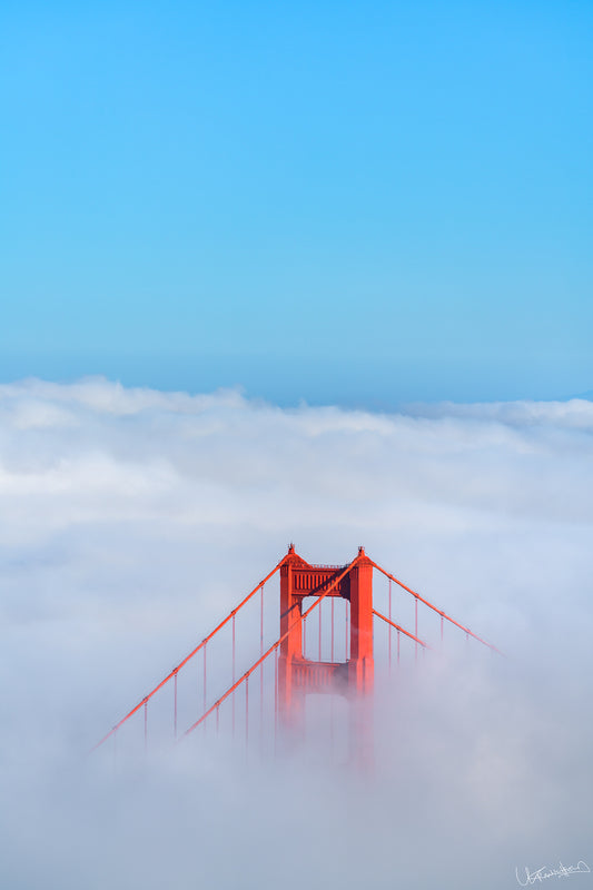 Red Golden Gate Bridge partially obscured by fog with a clear blue sky above.