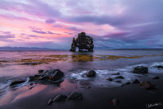 Stony beach with a large rock formation under a colorful sunset sky.