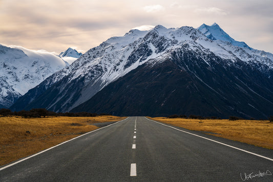 Long road leading to snow-capped mountains under a cloudy sky