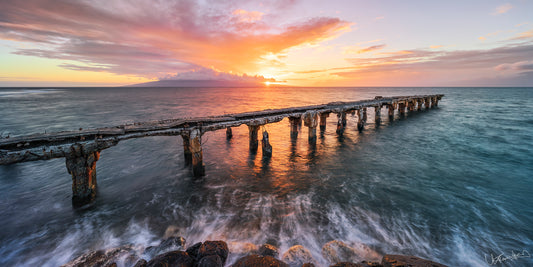 A photo depicting the broken down Mala ramp extending into the ocean, with the sea waves in motion and a sunset in the background. 