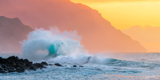 A photograph capturing a large wave with a heart crashing against a rocky shore at sunset with the Na Pali coast in the background.