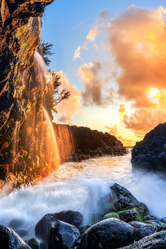 A scenic view of a waterfall at Queens Bath on Kauai, with the sun setting and casting warm shades on the rocks while the ocean waves crash below.