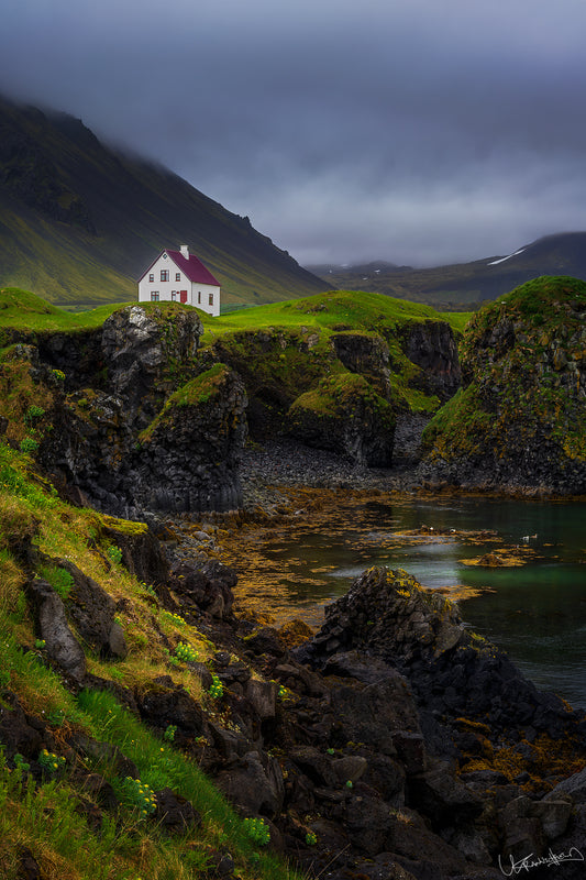 White house with a red roof on a cliff overlooking a body of water, surrounded by mountains under a dark sky.