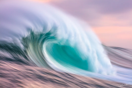 A soft long exposure image of a wave in the ocean, with a mix of blue and green colors and a hazy pink sky above.